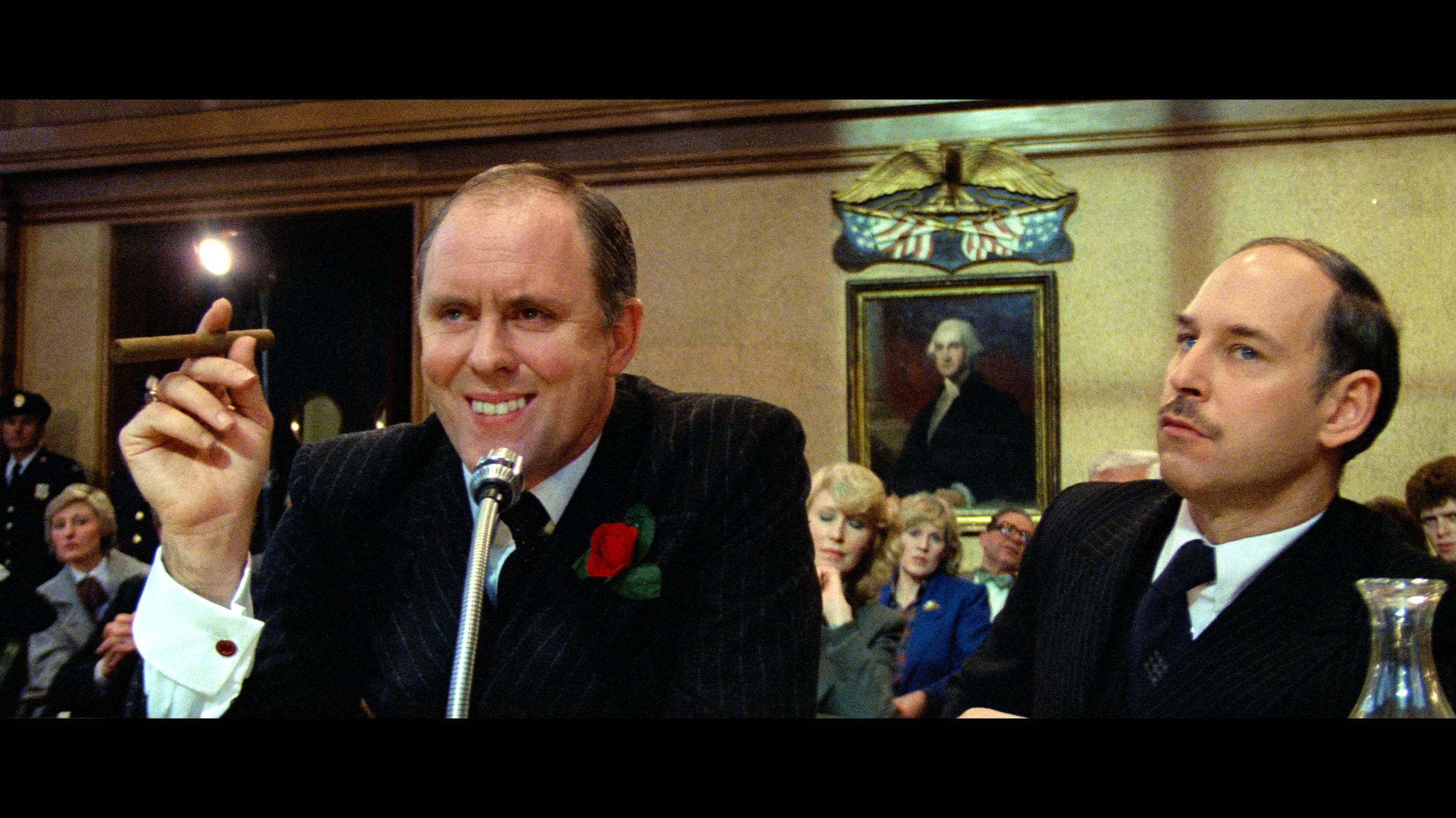 A man in a suit holding a cigar speaks into a microphone at a formal hearing, with people seated behind him and a portrait on the wall.