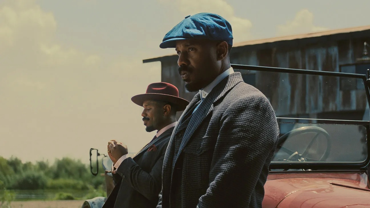Two men in vintage suits and hats stand beside a classic car in front of a rustic building, looking into the distance on a sunny day.