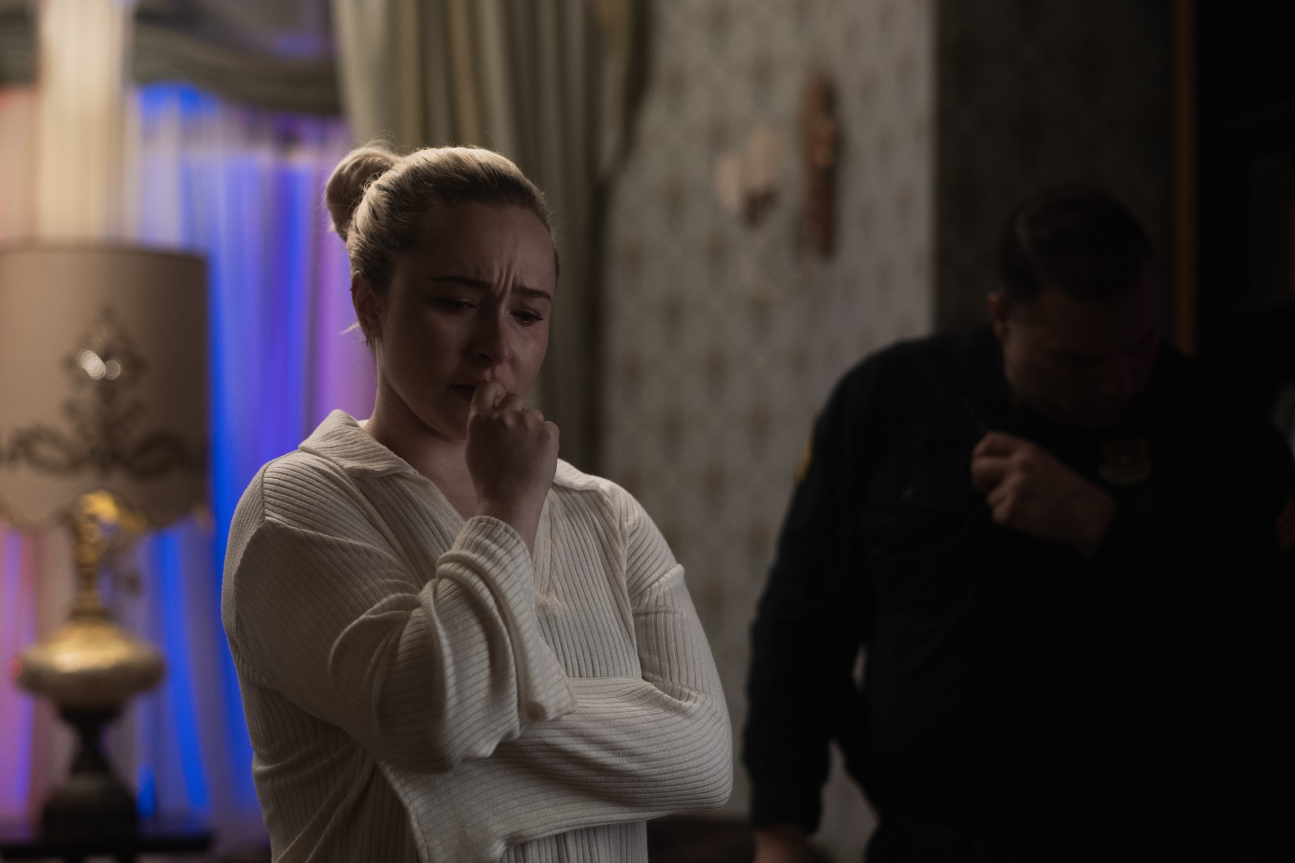 A woman stands with a worried expression and her hand near her mouth, while a police officer is seen in the background in a dimly lit room.