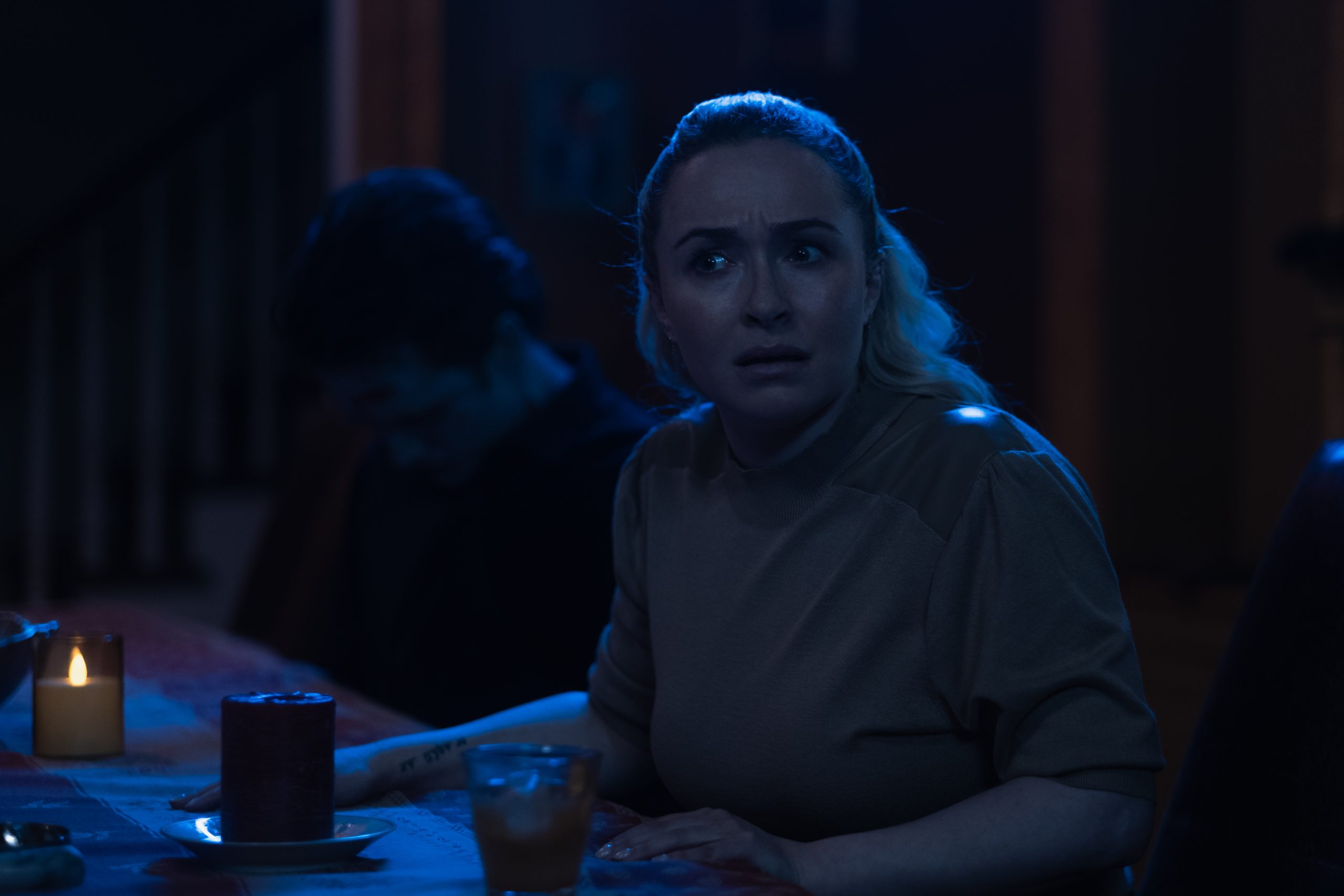 A woman sits at a dimly lit table looking concerned, while a man beside her sits slumped over. Lit candles and a glass are on the table.