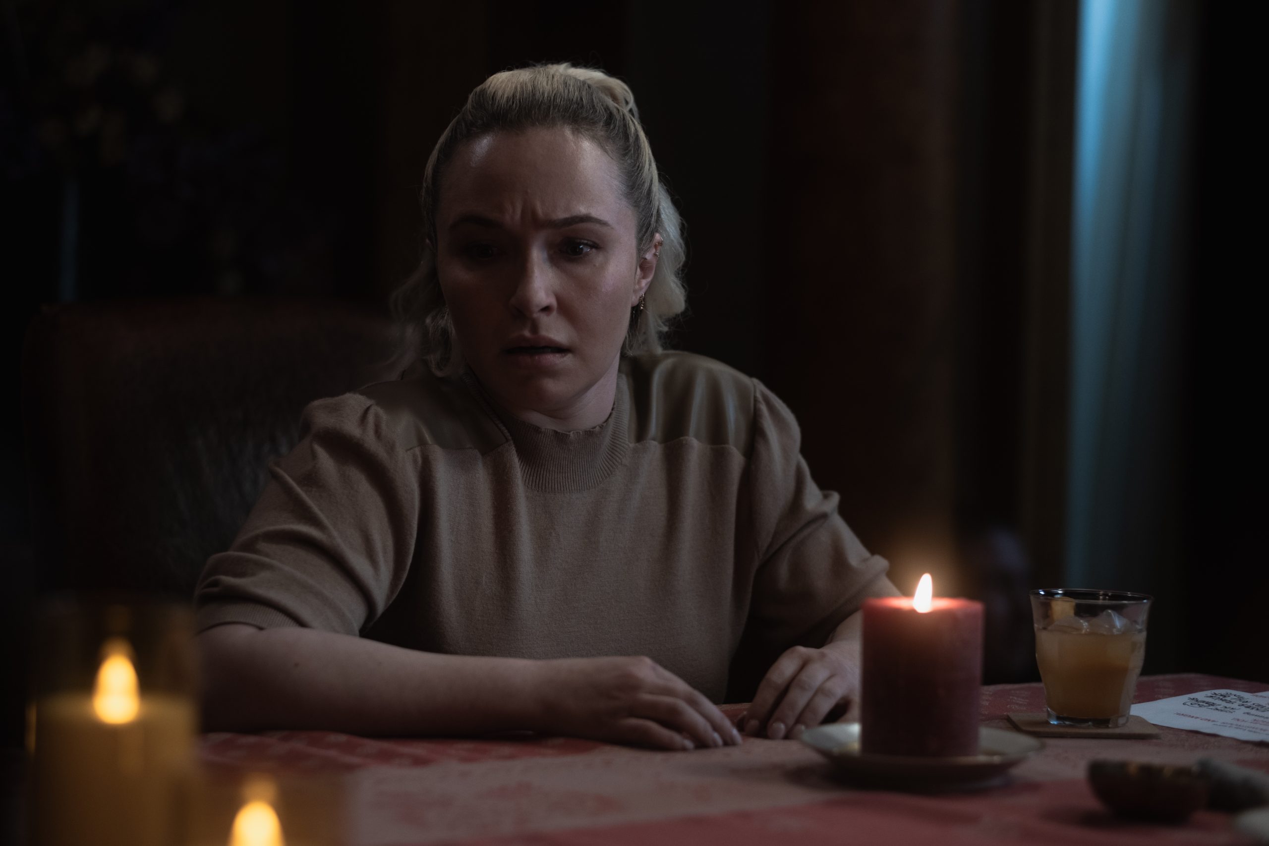 A woman with blonde hair sits at a dimly lit table with a red candle, drink, and paper, looking concerned.