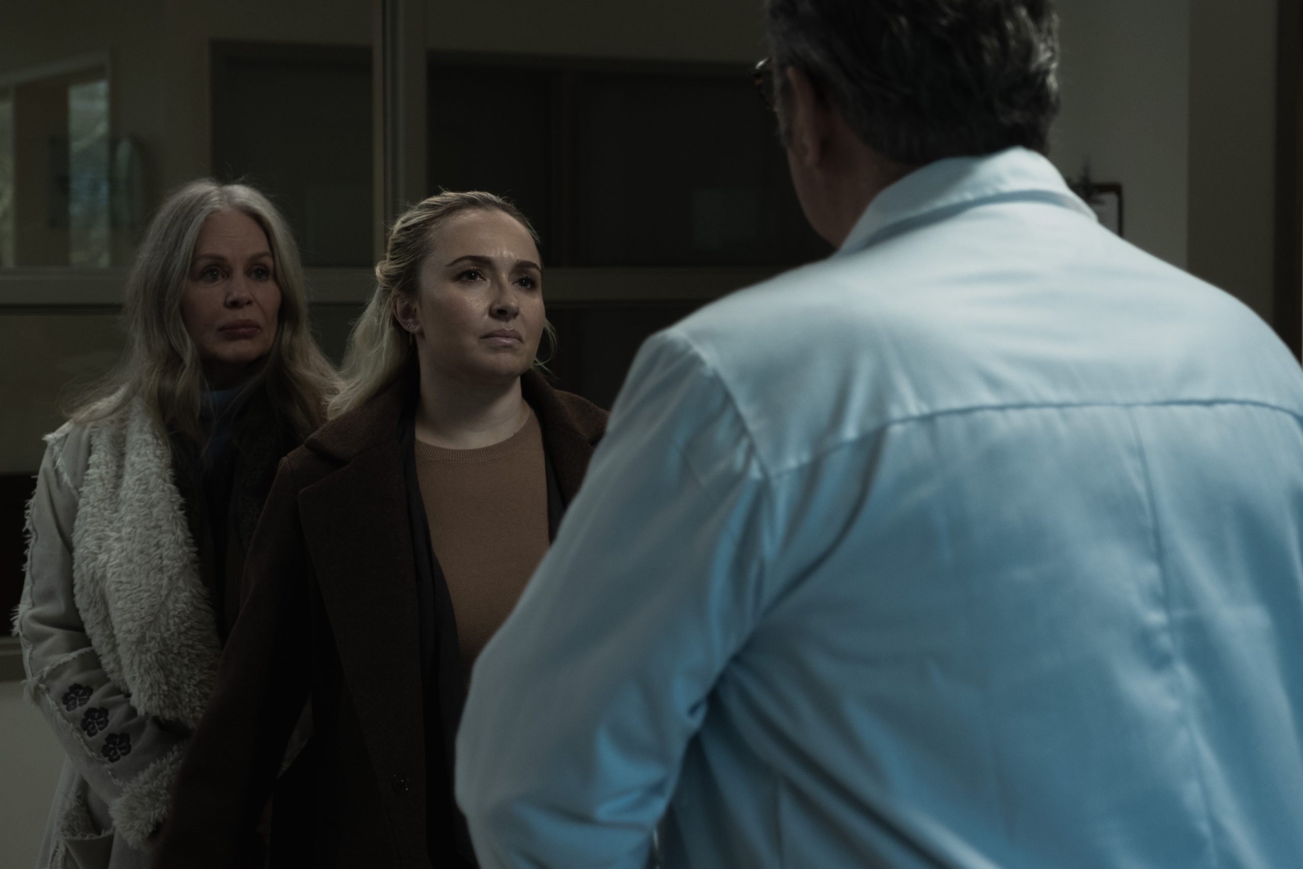 Two women stand facing a man in a white coat inside a dimly lit room, with serious expressions on their faces.