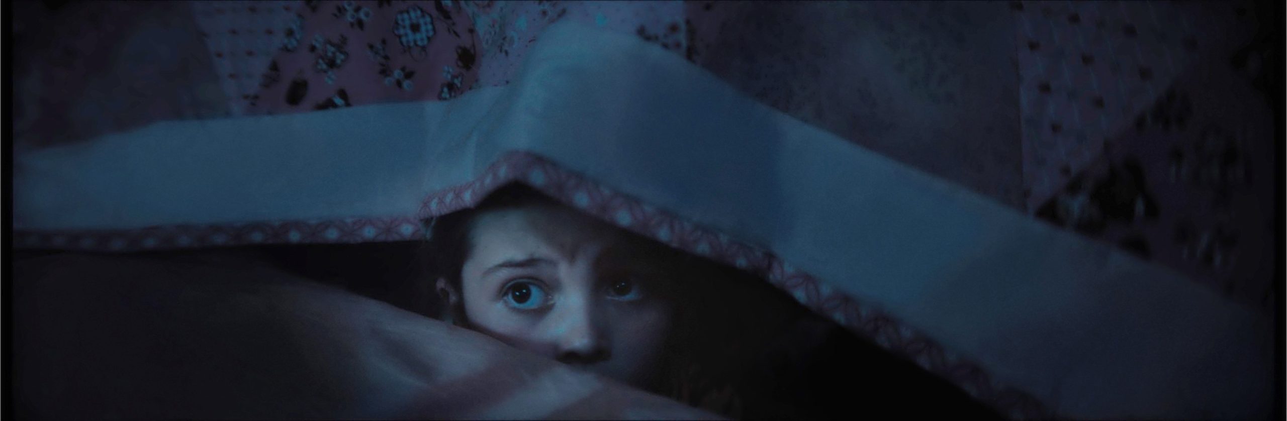 A child with wide eyes looks up while hiding under patterned blankets in dim lighting.