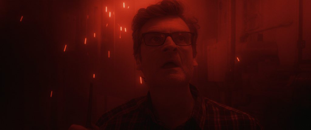 A man wearing glasses stands in a dimly lit room illuminated by red light, with sparks flying around him.