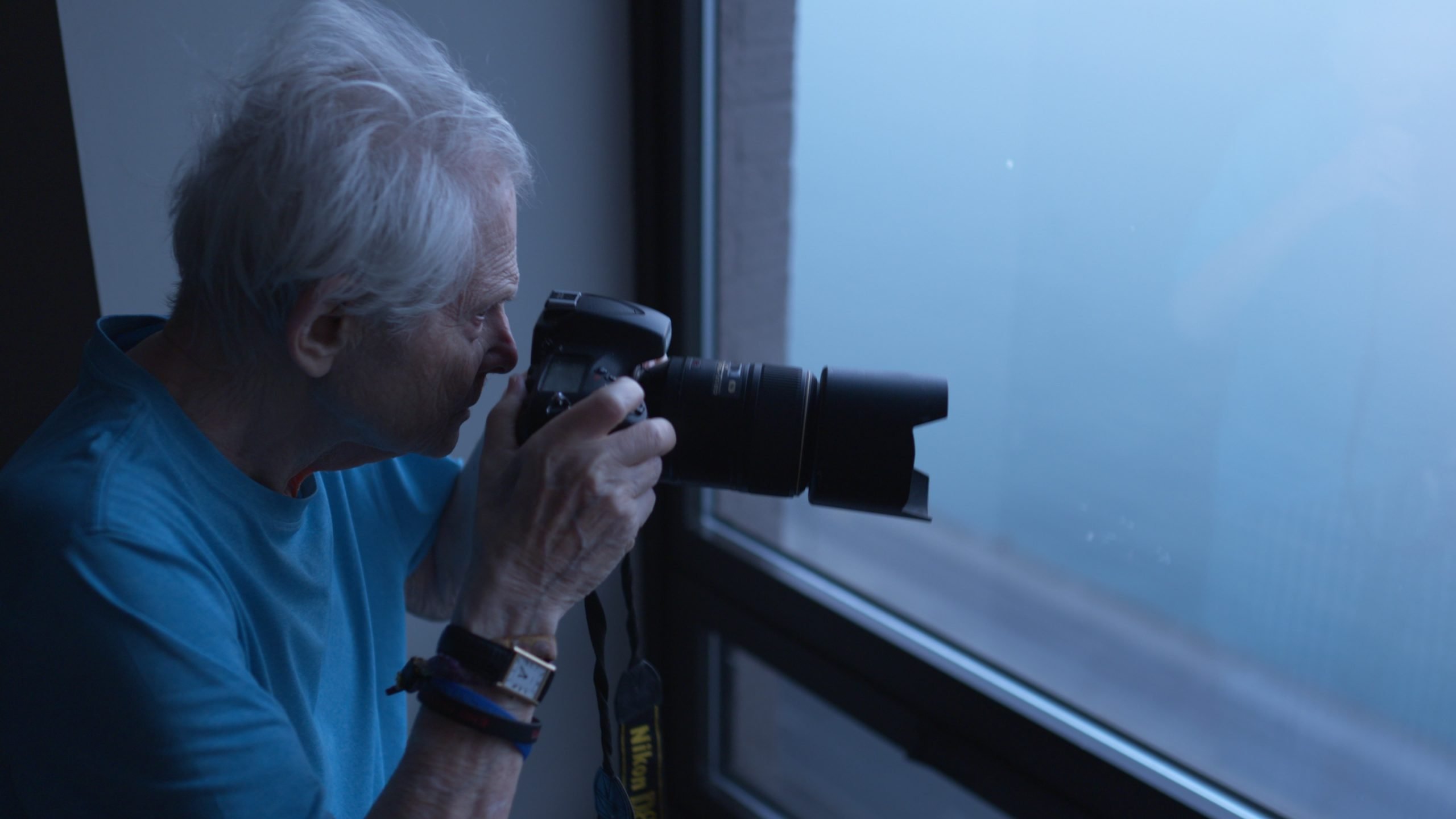 An elderly person with white hair holds a DSLR camera, aiming it out of a window on a cloudy day.