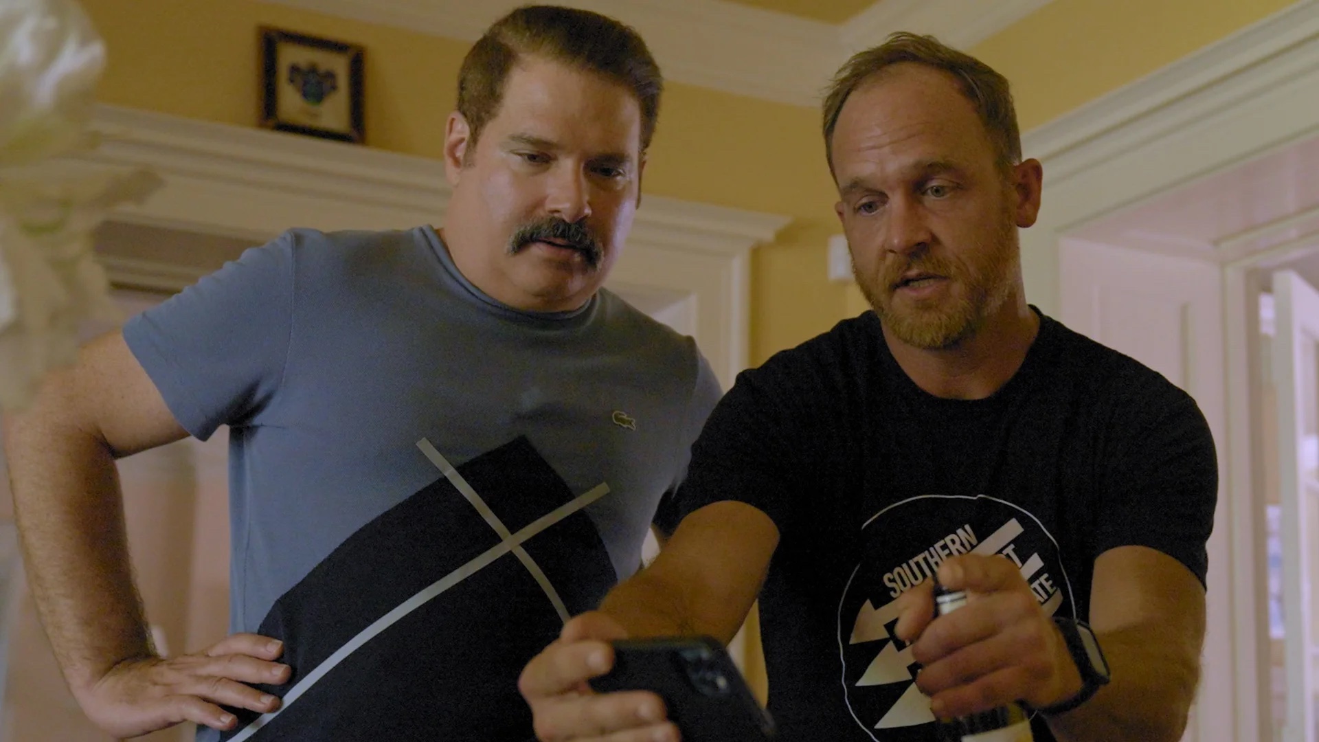 Two men standing indoors look at a smartphone, with one holding the phone and gesturing as they focus on the screen.