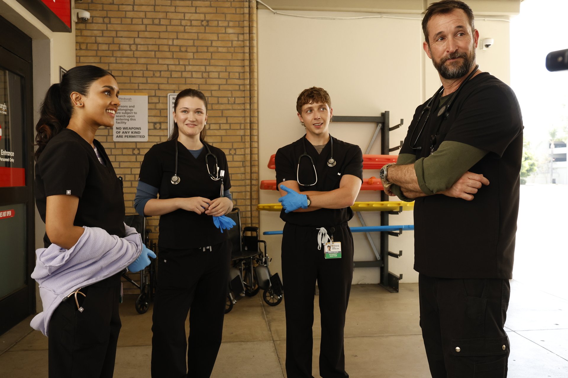 Four medical professionals in black scrubs stand outside a building, engaged in conversation. Three are smiling, and one is looking off to the side with arms crossed. Medical equipment is visible behind them.