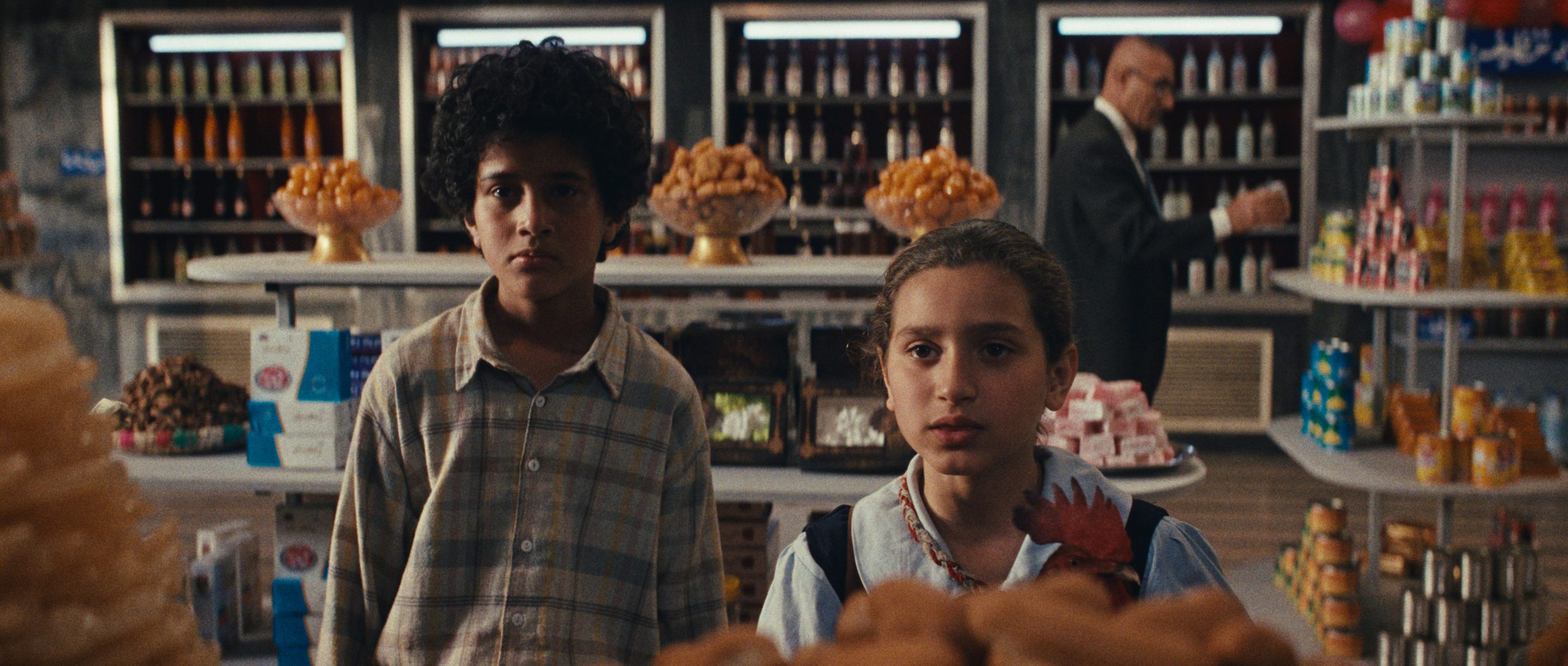 Two children stand in a grocery store, looking ahead; shelves stocked with goods and a man in a suit are visible in the background.