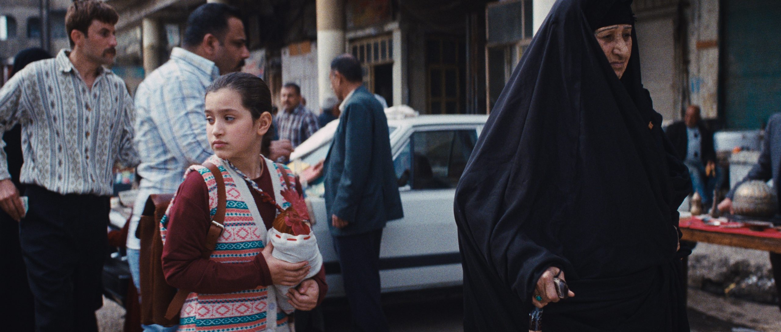 A young girl with a backpack holds a wrapped bundle, standing next to a woman in a black abaya on a busy street with people and cars in the background.