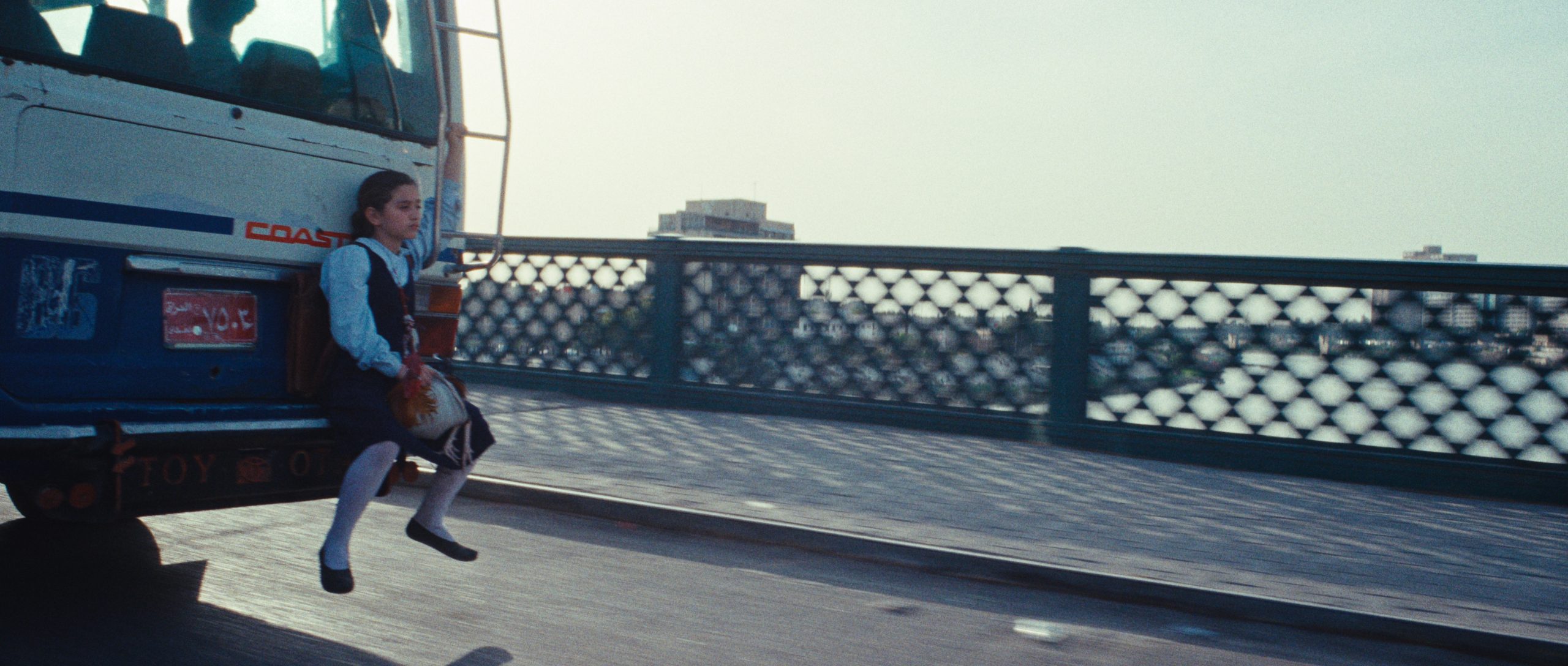 A young girl in a school uniform rides on the rear bumper of a moving bus over a bridge with a river and city buildings in the background.