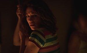 A woman with wavy hair and a striped shirt looks over her shoulder with a concerned expression in a dimly lit setting.