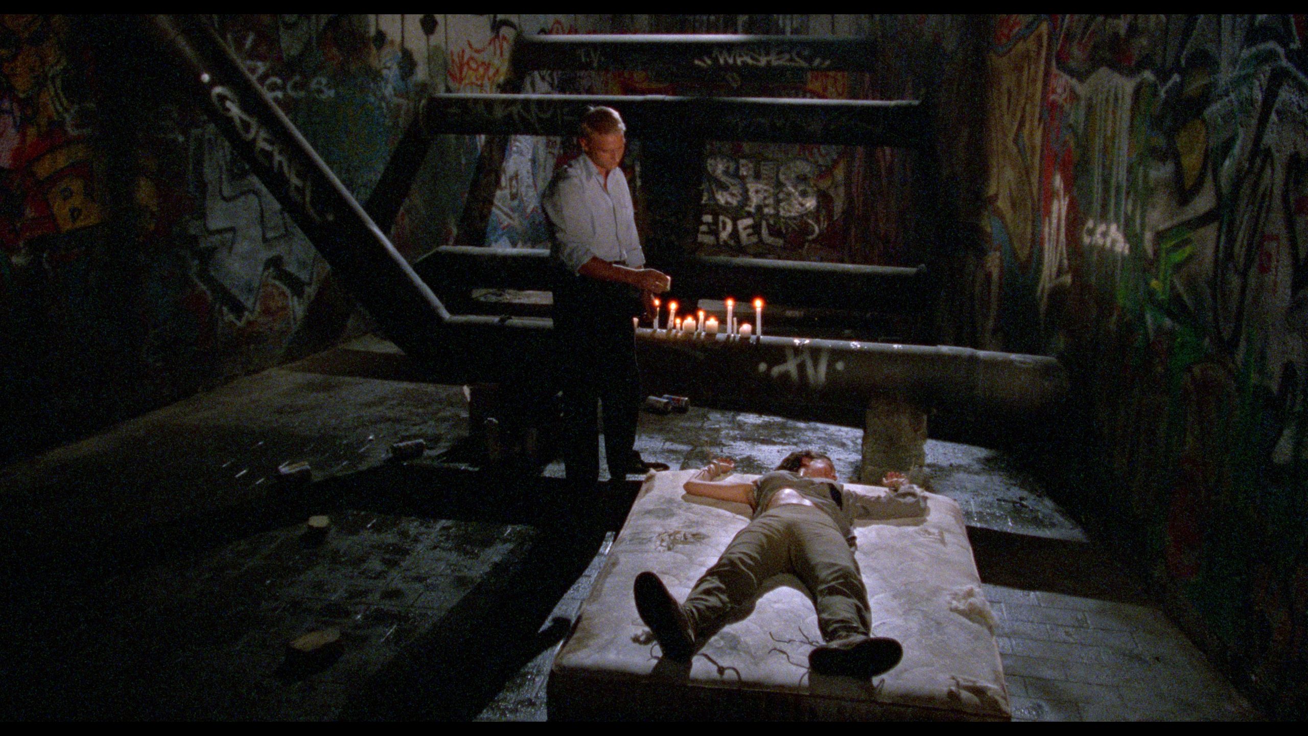 A man stands near lit candles beside a person lying on a mattress under a large metal staircase in a graffiti-covered, dimly lit room.
