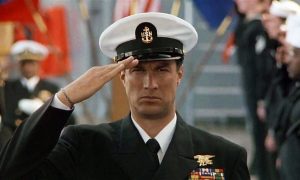 A man in a U.S. Navy dress uniform salutes while standing outdoors, with military personnel and flags in the background.