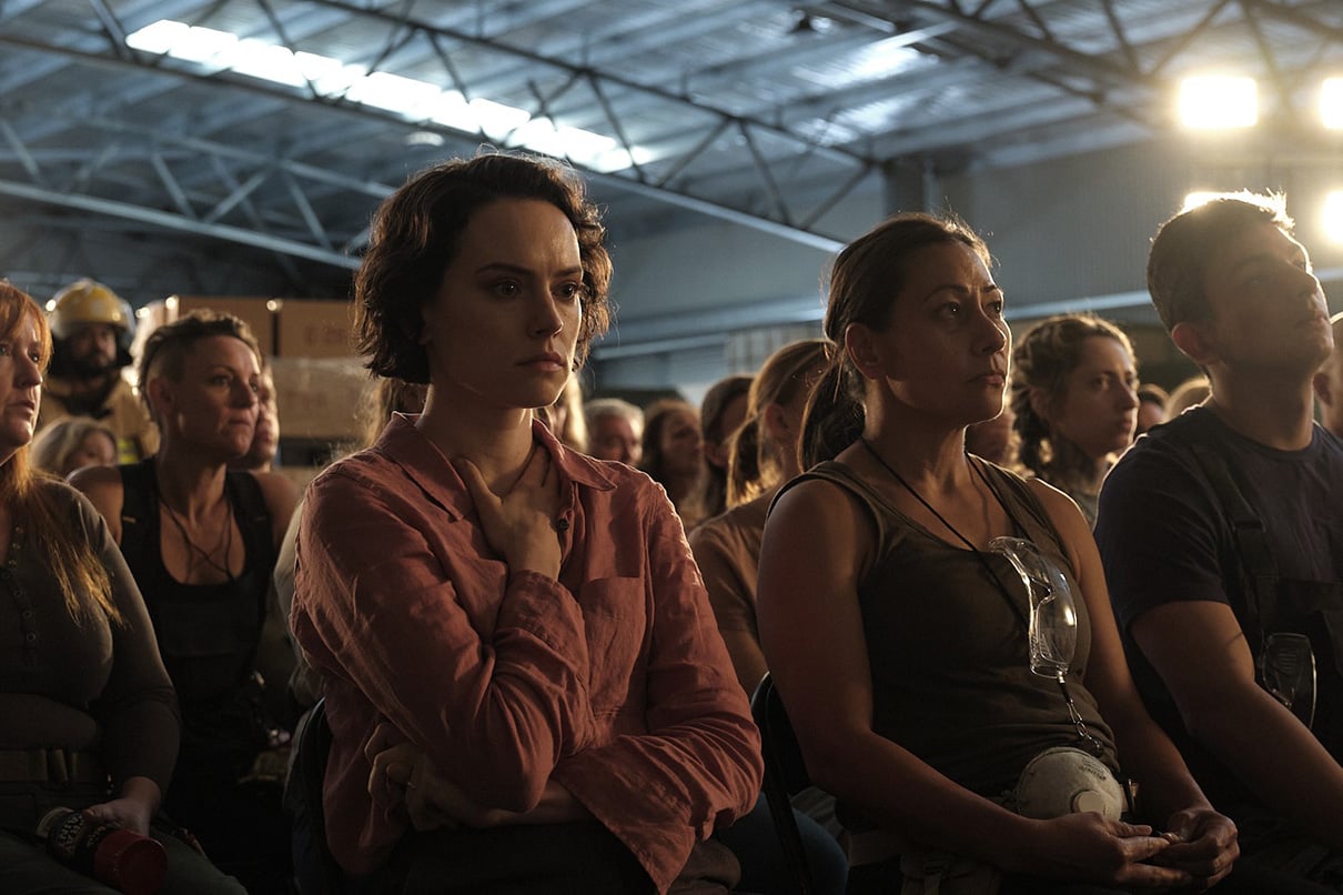 A group of people sit closely together in a warehouse, attentively listening to someone off-camera. The lighting is bright in the background.