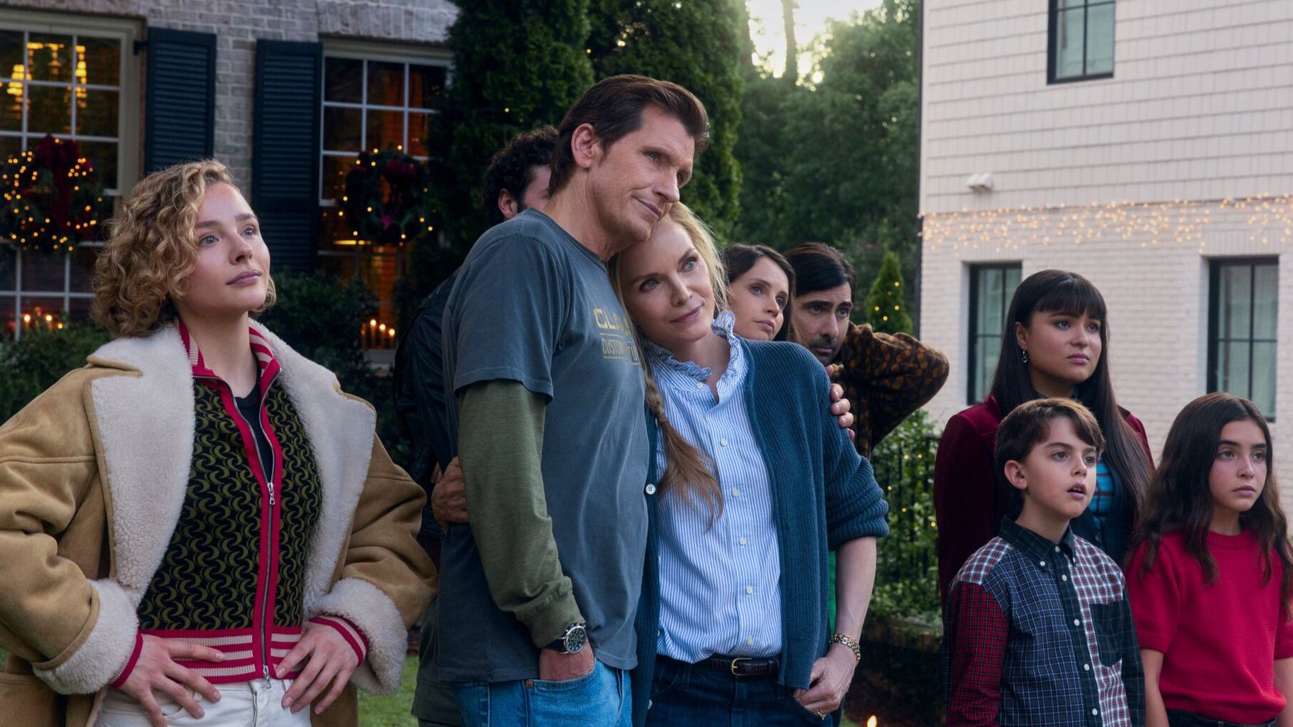 A group of people, including adults and children, stand close together outdoors in front of decorated houses, looking in the same direction with neutral and thoughtful expressions.