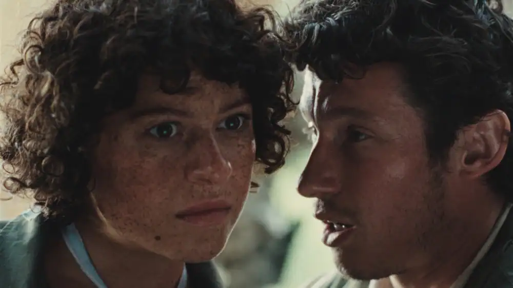 A person with curly hair and freckles looks intently ahead while another person speaks closely beside them, both displaying serious expressions.