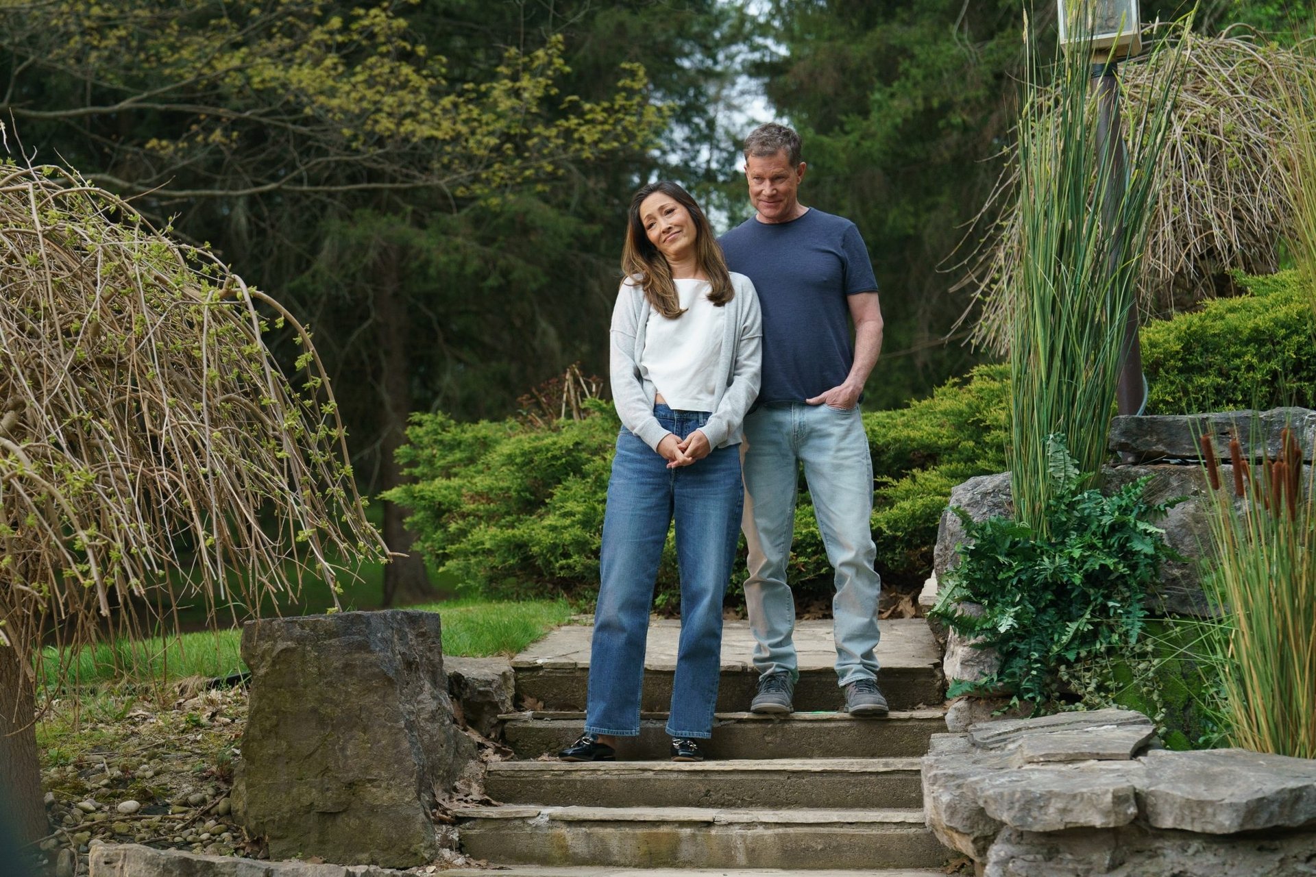 A woman and a man stand together on stone steps in a garden surrounded by greenery and trees.