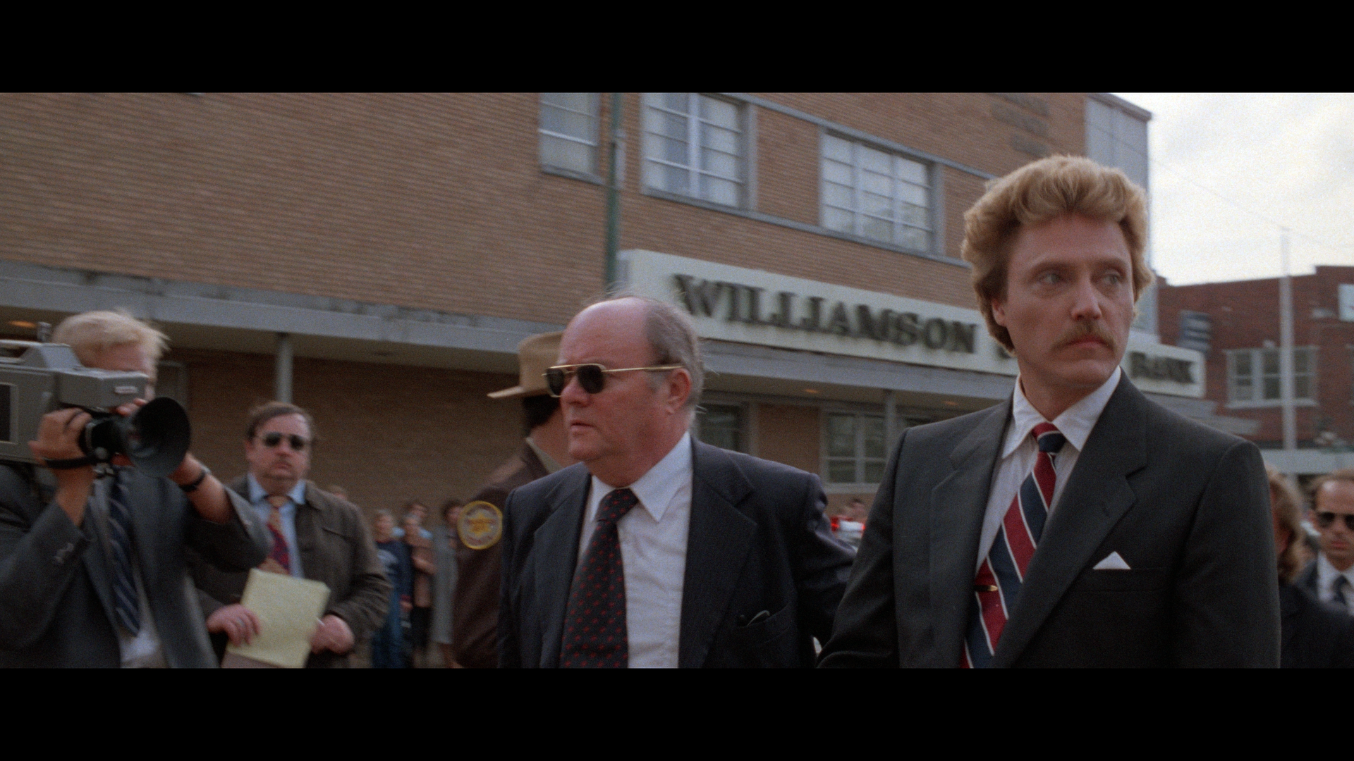 Two men in suits walk outside a brick building labeled "Williamson" as a film crew and police officers stand nearby.