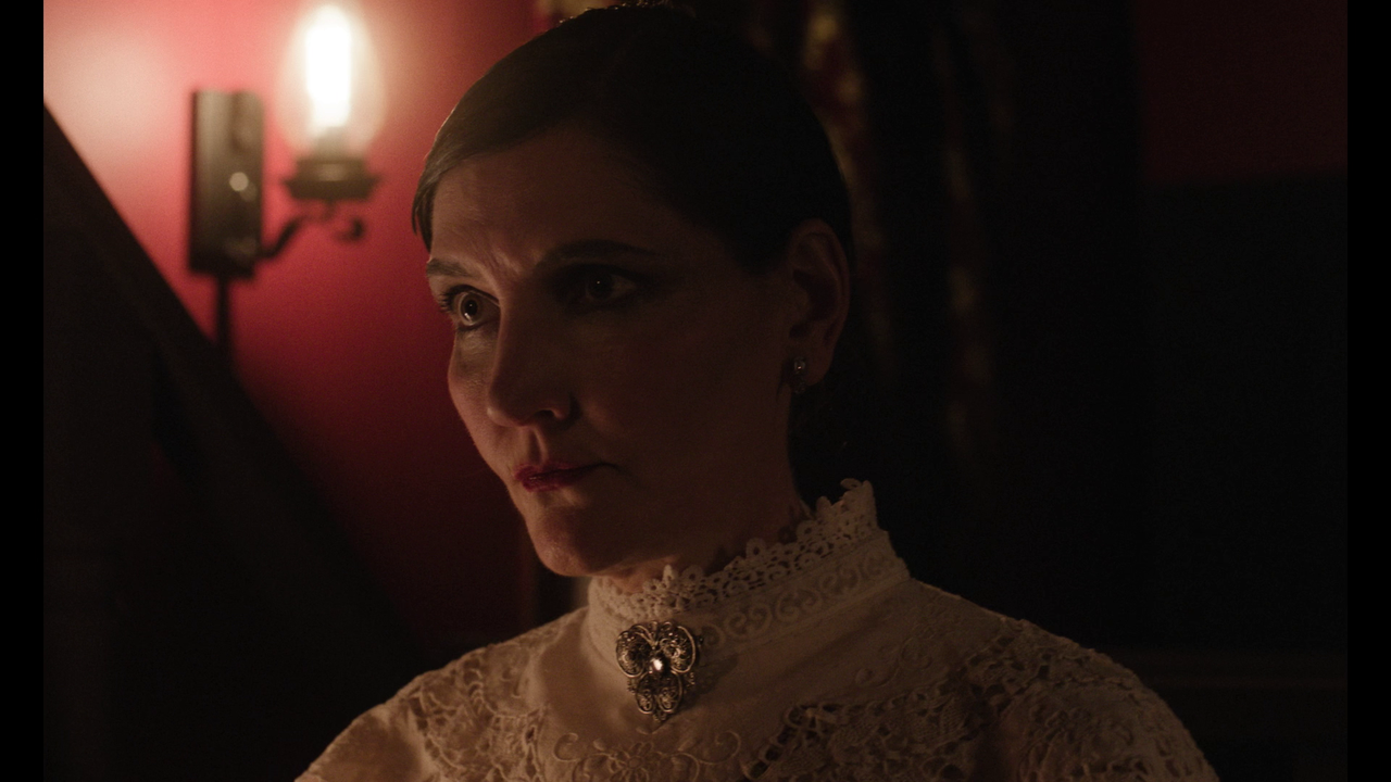 A woman in a high-collared lace dress sits indoors under warm lighting, with a decorative brooch at her neck and a sconce light on the red wall behind her.