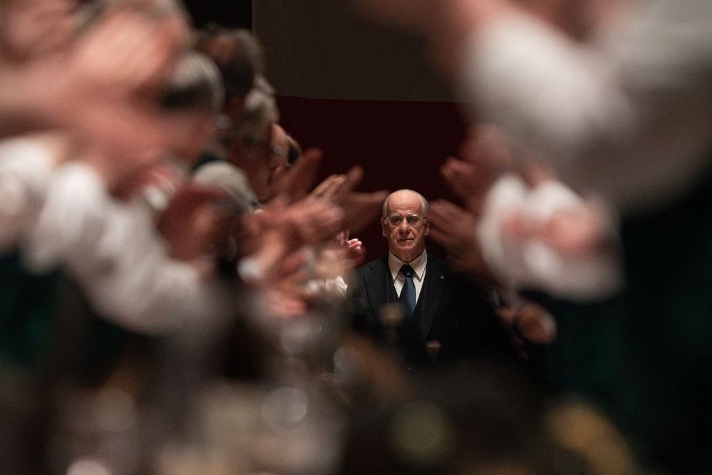 A man in a suit stands in focus at the center of a room, with blurred people on both sides clapping.