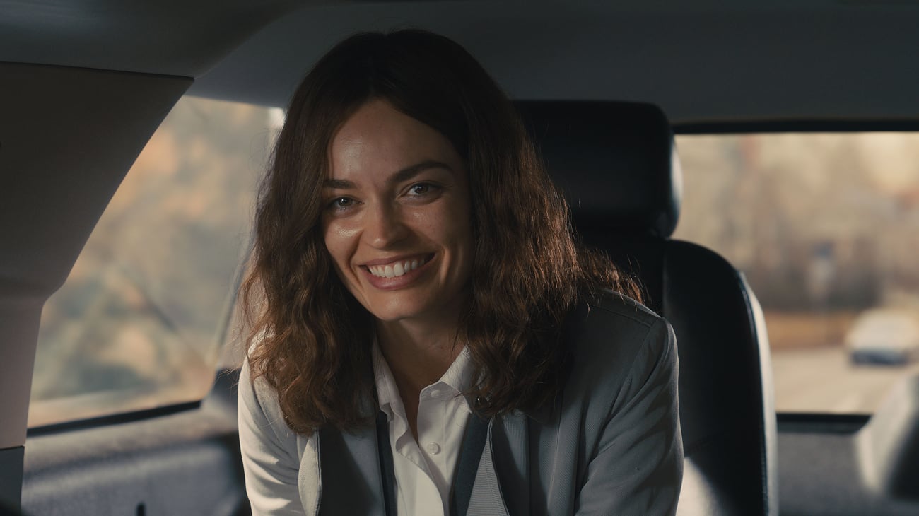 A woman with wavy brown hair, wearing a grey blazer and white shirt, smiles while sitting in the back seat of a car.