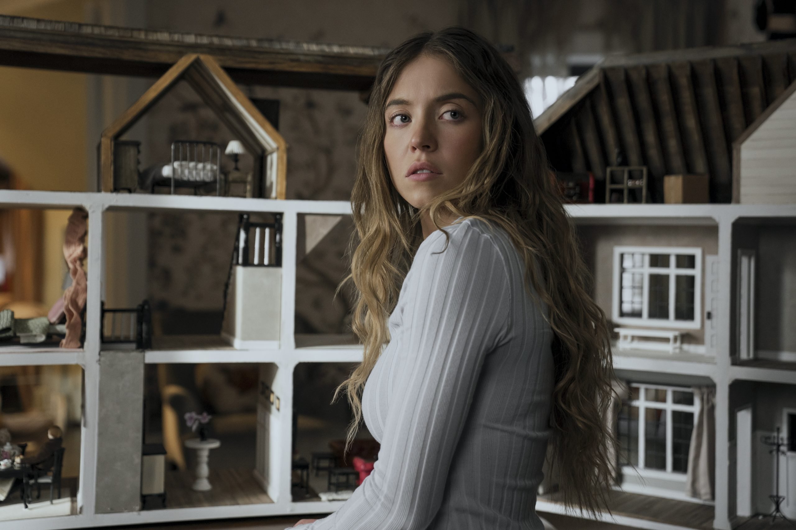 A young woman with long wavy hair in a white ribbed top sits in front of a large, multi-level dollhouse, looking to her left with a neutral expression.
