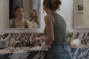 A woman cleans a bathroom sink while looking into a mirror that reflects another woman standing behind her.