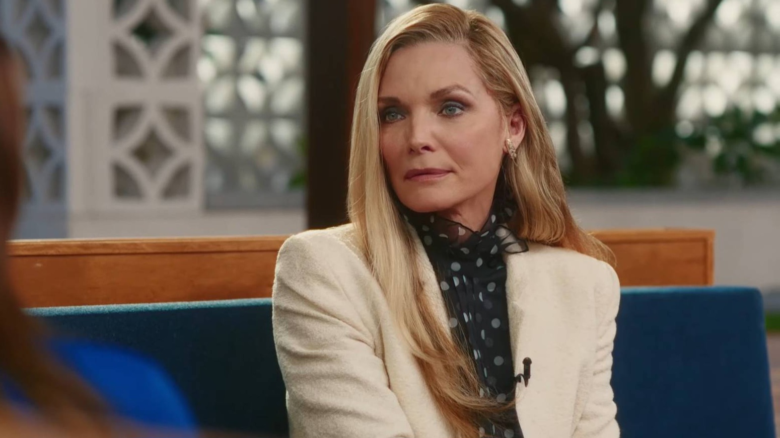 A woman with long blonde hair wearing a white blazer sits on a blue bench indoors, looking thoughtfully to her left.