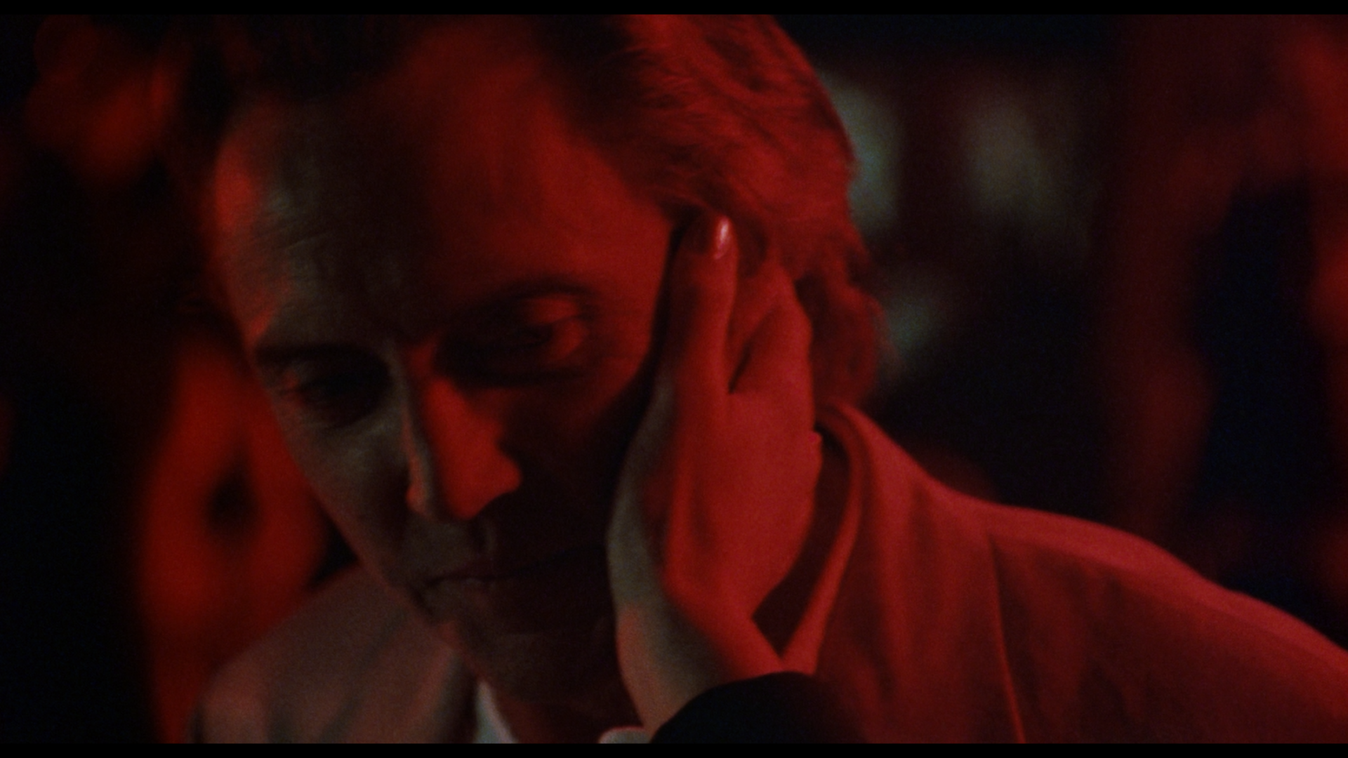 A person in a white jacket is gently touched on the cheek by a hand, with red lighting casting dramatic shadows on their face.