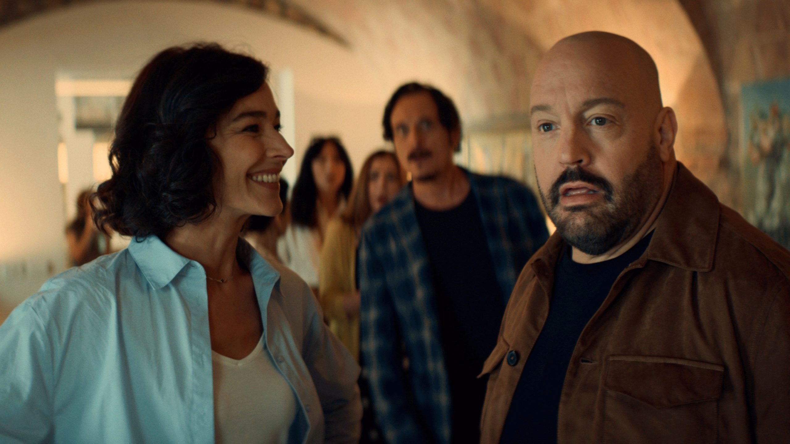 Un grupo de adultos está en el interior, con una mujer con una camisa azul claro sonriendo y un hombre con una chaqueta marrón que parece sorprendido en primer plano.