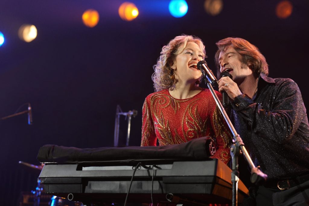 A woman in a red dress plays keyboard while singing with a man in a black shirt on stage under colorful lights.