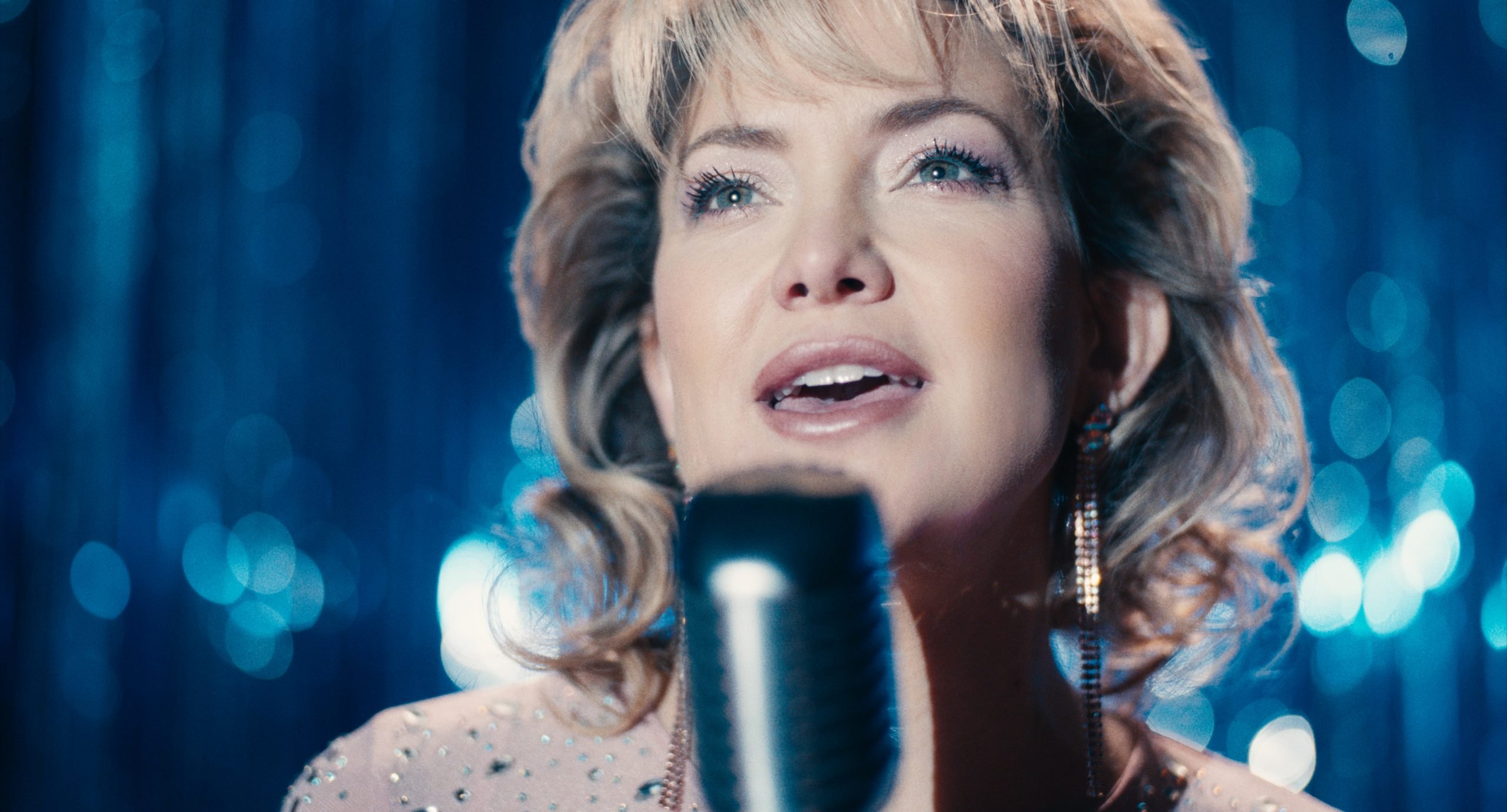 A woman with blond hair sings into a vintage microphone on stage, with blue sparkling curtains in the background.