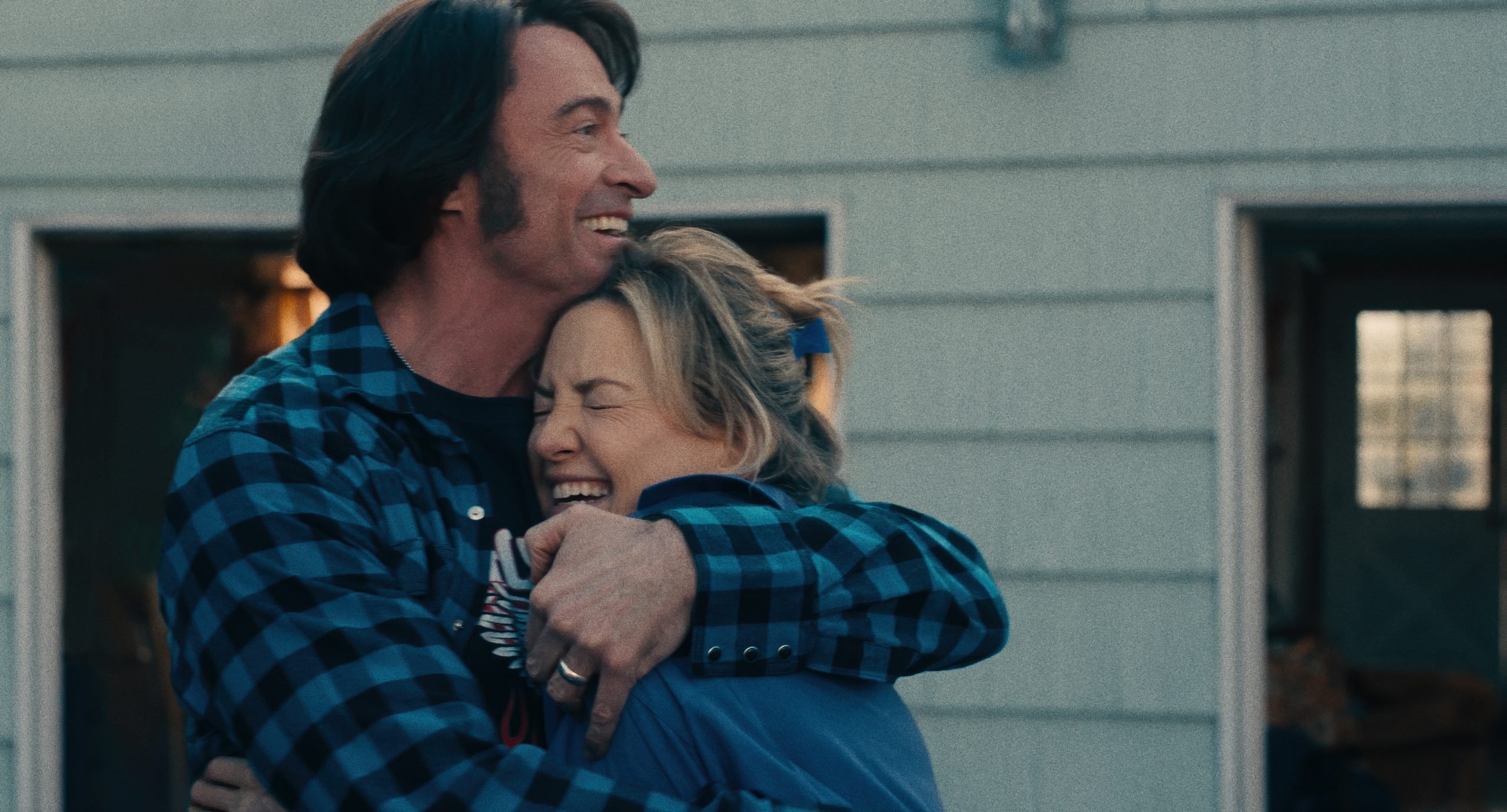 A man in a blue plaid shirt hugs a woman, both smiling, outside a building with two open garage doors.