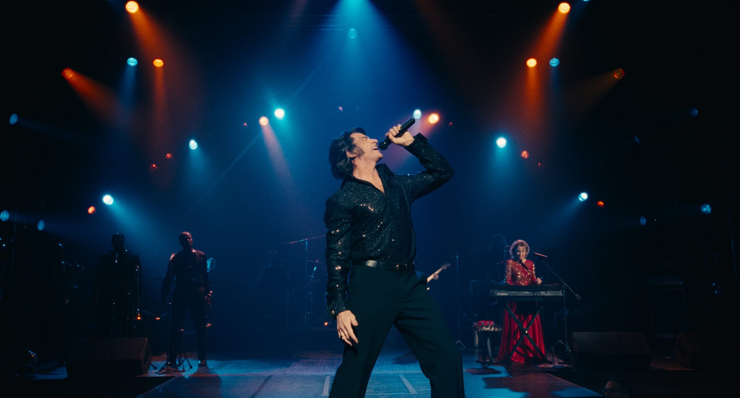 A man in a sparkling shirt sings passionately into a microphone on stage, with musicians and colorful stage lights in the background.