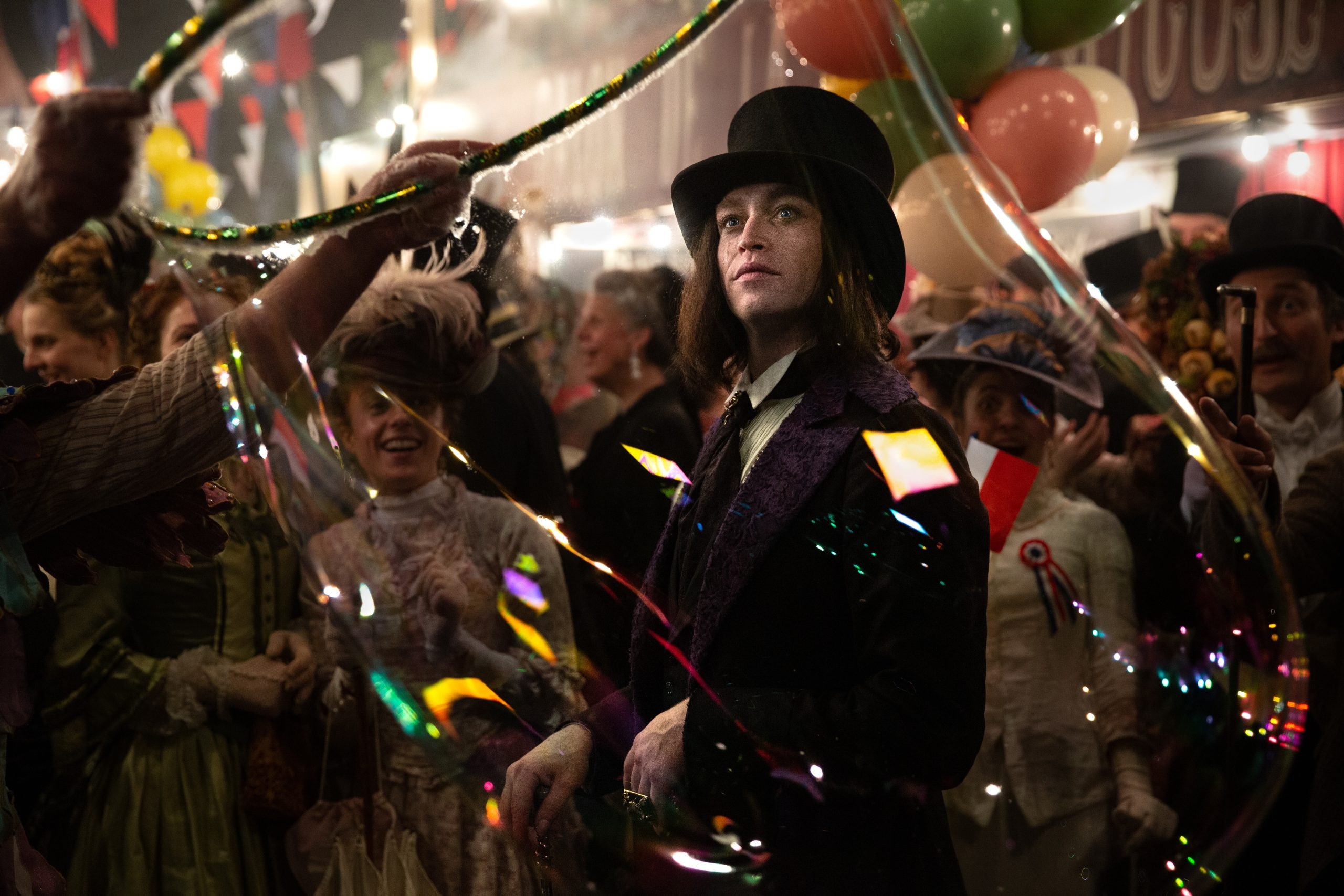A man in a top hat and coat stands amid a lively, crowded carnival scene, surrounded by people in period costumes and large soap bubbles.