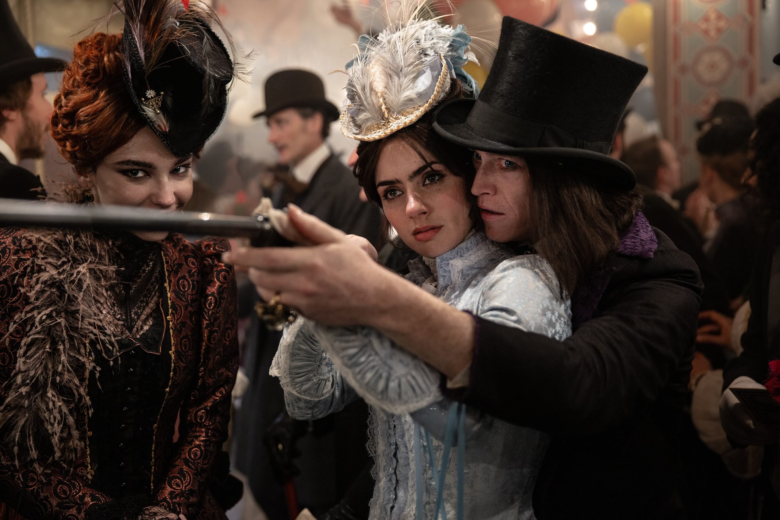 Three people in elaborate Victorian-style costumes; one woman aims a rifle with guidance from a man in a top hat while another woman in dark attire watches closely.