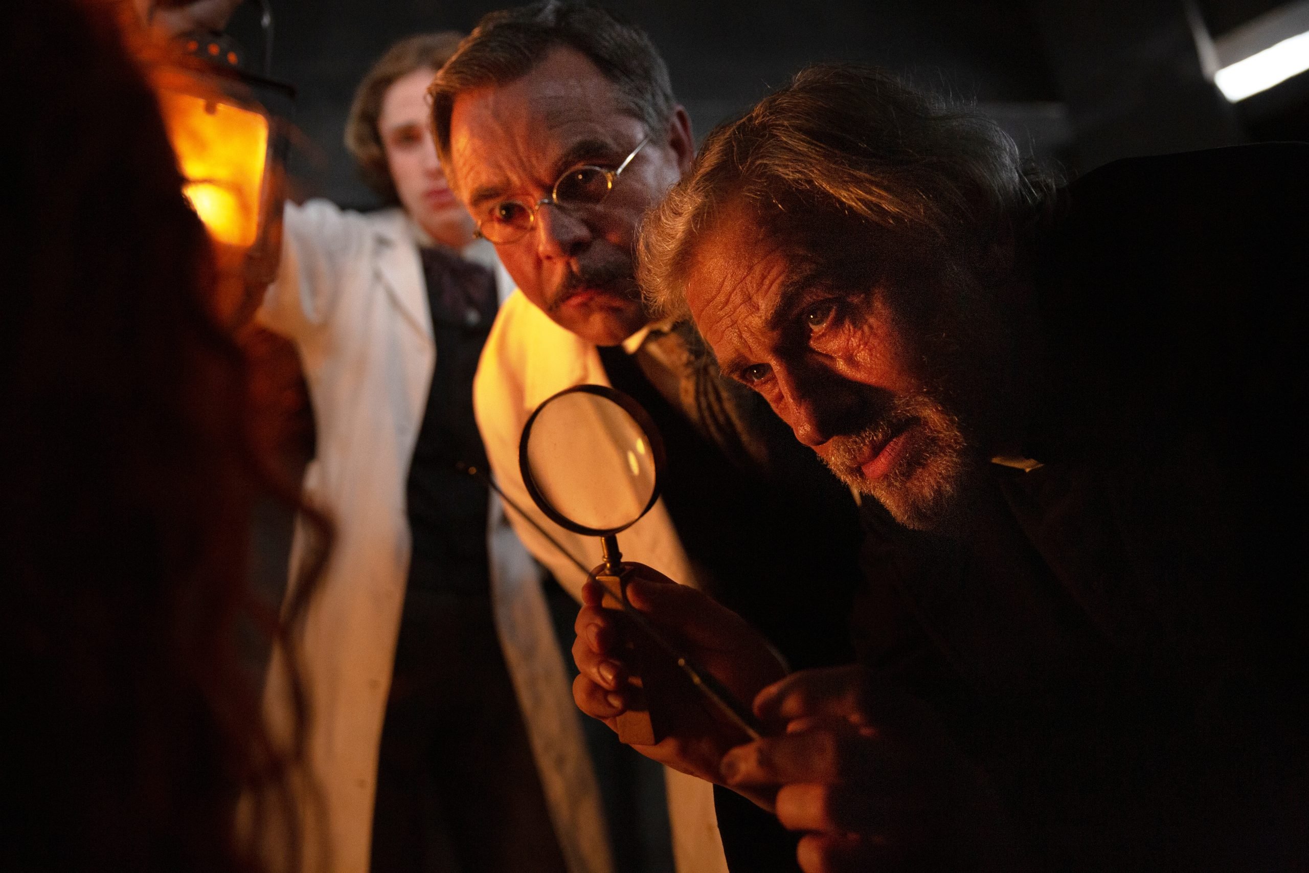 Two men in lab coats examine something closely with a magnifying glass and lantern in a dimly lit room, while a third person stands in the background.