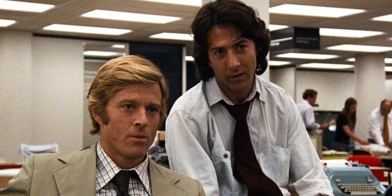 Two men in dress shirts and ties sit in a busy office with fluorescent lighting, looking intently ahead. Other people are working in the background.