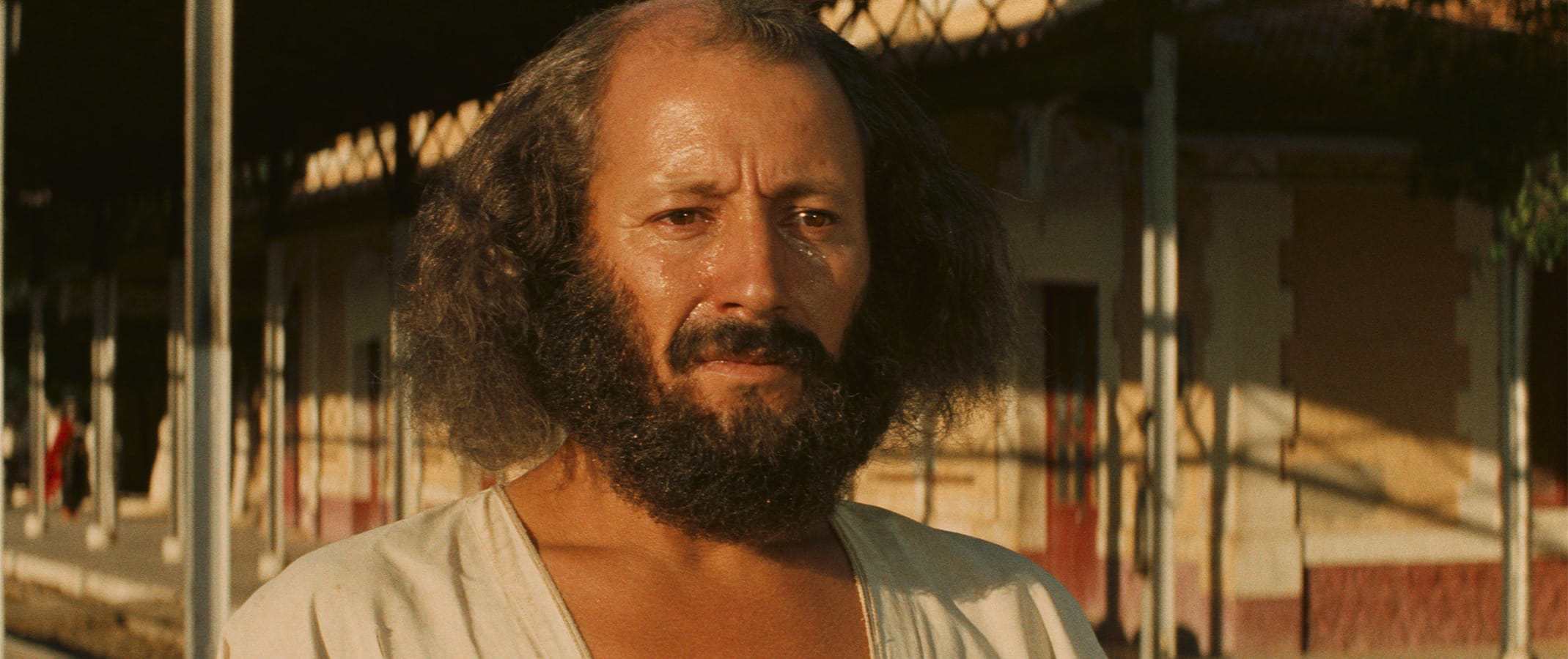 A man with a beard and shoulder-length hair stands outdoors near a building, looking slightly downcast in warm sunlight.