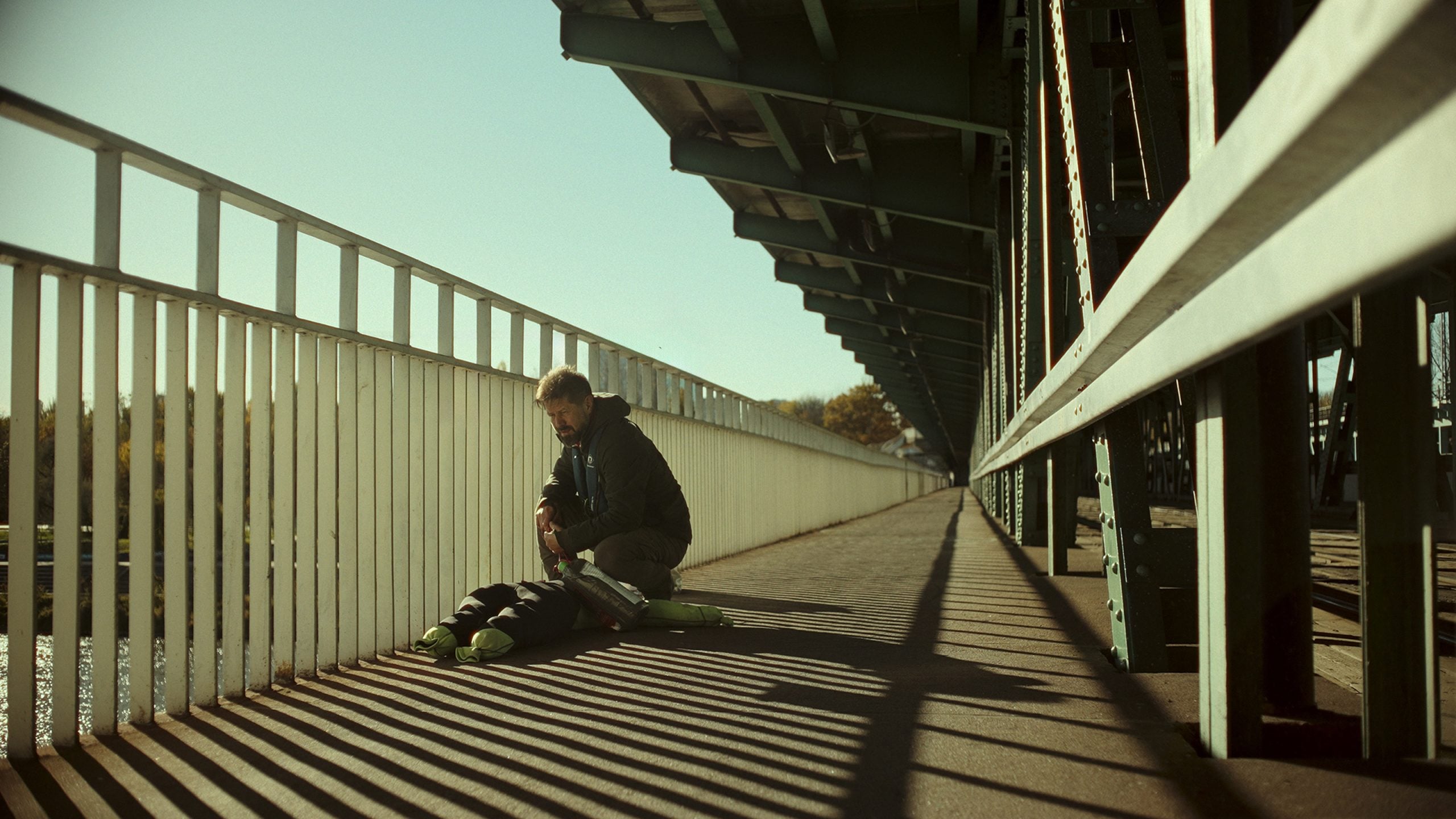 A man kneels beside a person lying on the ground on a pedestrian bridge, with sunlight casting shadows from the railings.