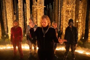 A group of five people stand in a circle of firelight, with tall carved wooden pillars and burning candles in the background at night.