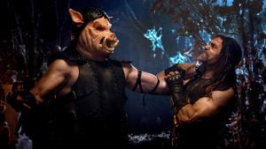 A man in armor with a pig mask grabs another man by the neck in a dark, fantasy-themed forest setting.