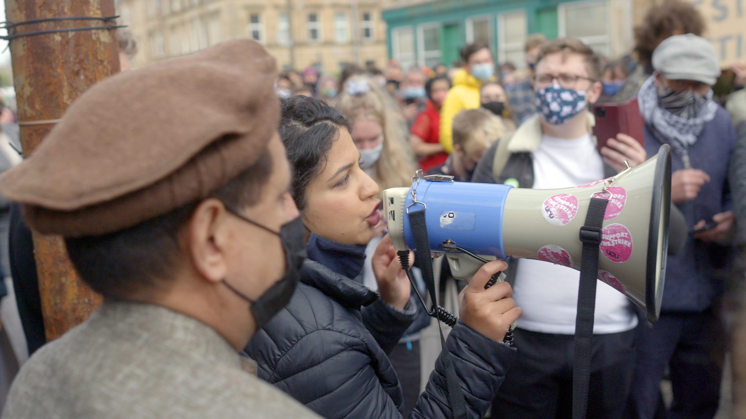 A woman speaks into a megaphone surrounded by a crowd of people, some wearing masks and holding phones, at an outdoor gathering or protest.