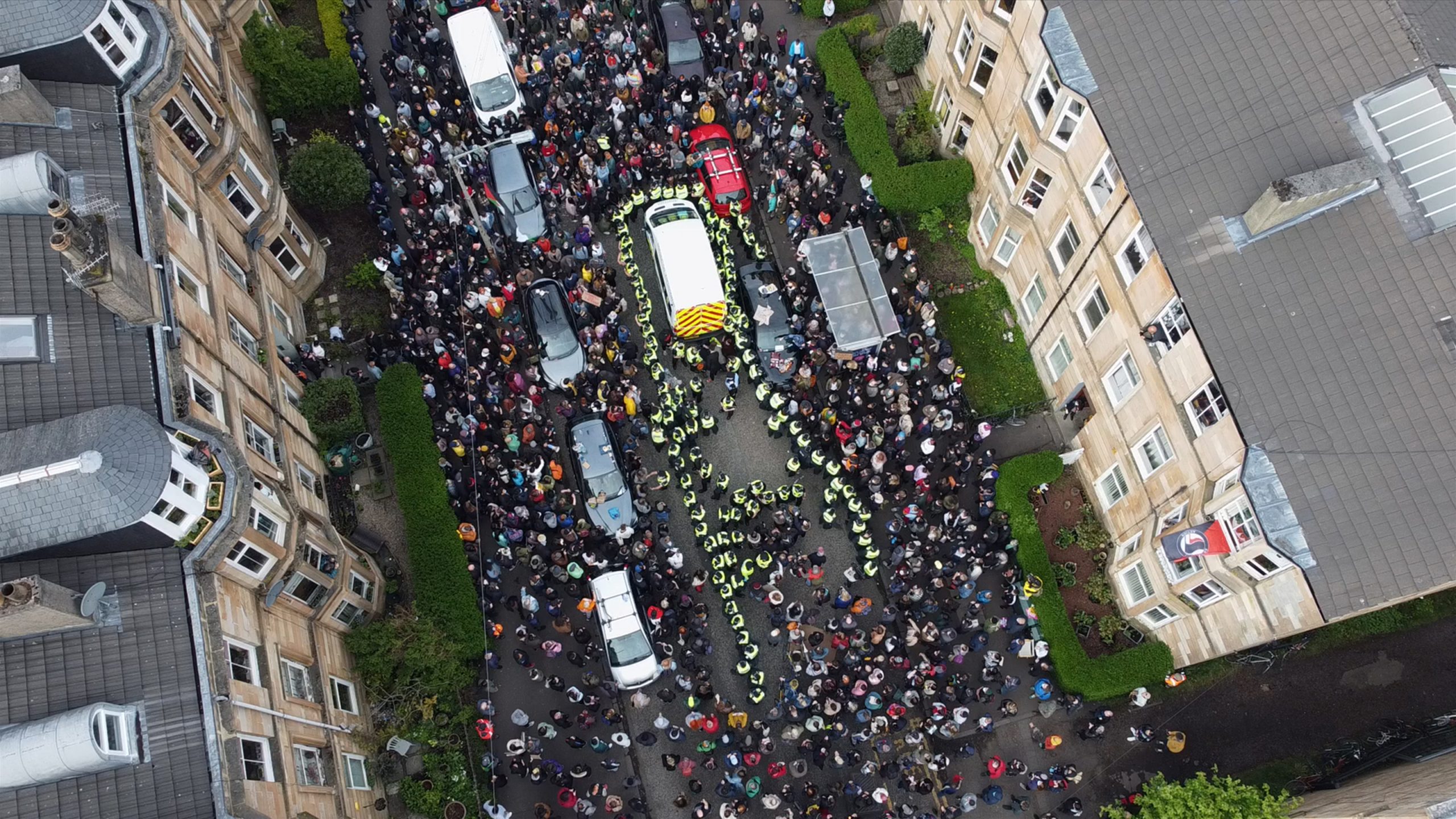 Aerial view of a crowded street with police surrounding a white vehicle, emergency vehicles, and people gathered between buildings.