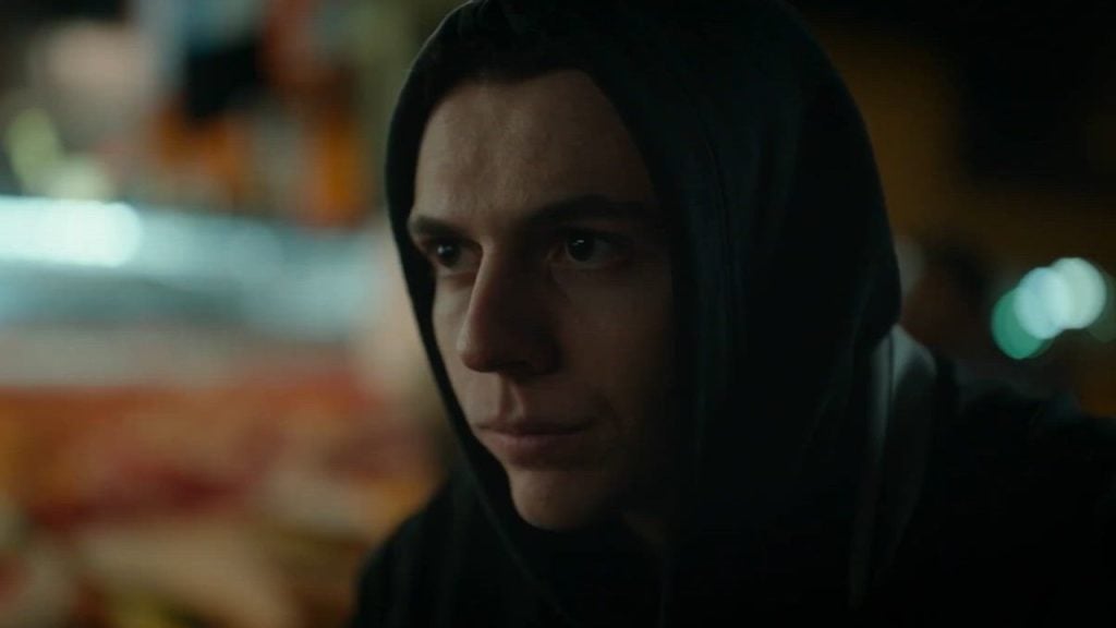 A person wearing a dark hoodie looks ahead with a serious expression in a dimly lit setting.