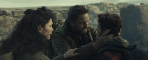 A man holds a boy’s face with both hands while a woman stands nearby; they are outdoors in a rocky, mountainous landscape.