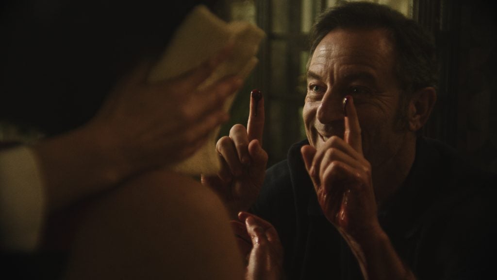 A man with blood on his fingers smiles while holding his hands up, facing a person whose face is partially covered by a cloth.