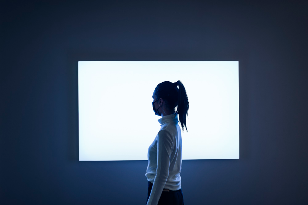A person in a white turtleneck and face mask stands in profile before a large, bright rectangular light on a dark wall.