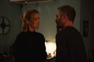 A woman and a man stand facing each other indoors, engaged in a serious conversation in a dimly lit room.