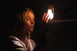 A woman in a dimly lit room reaches toward a glowing light bulb, her face illuminated by its light.